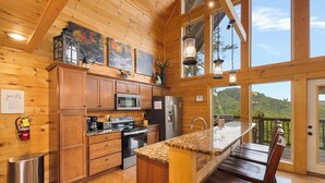 Une cuisine de style rustique, avec des murs en bois, un grand îlot central, des armoires en bois et une vue sur une colline boisée.