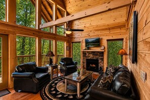 Interior de una cabaña de madera con chimenea, muebles de cuero y ventanas amplias.