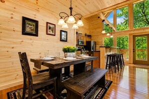 A wooden interior with a dining table set for four, kitchen cabinets, and a large window with a view of trees. - Cabin + FREE Resort Amenities | Pigeon Forge (Pigeon Forge)