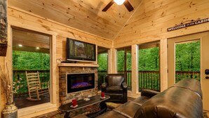 Interior de una cabaña con chimenea de piedra, asientos de cuero, un televisor y un letrero que dice 'Welcome to the Cabin'.