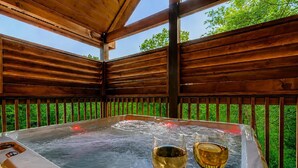 Un jacuzzi con dos copas de vino, rodeado de barandas de madera y follaje.