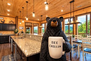 A bear mascot holding a mug with 'BEE CHILLIN'' text in a wooden cabin-style kitchen with granite countertops and large windows. - Near Dollywood + Resort Amenities Included (Sevierville)