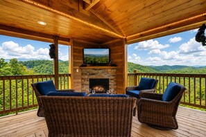 A wooden deck with wicker furniture and a stone fireplace, overlooking a forested landscape.