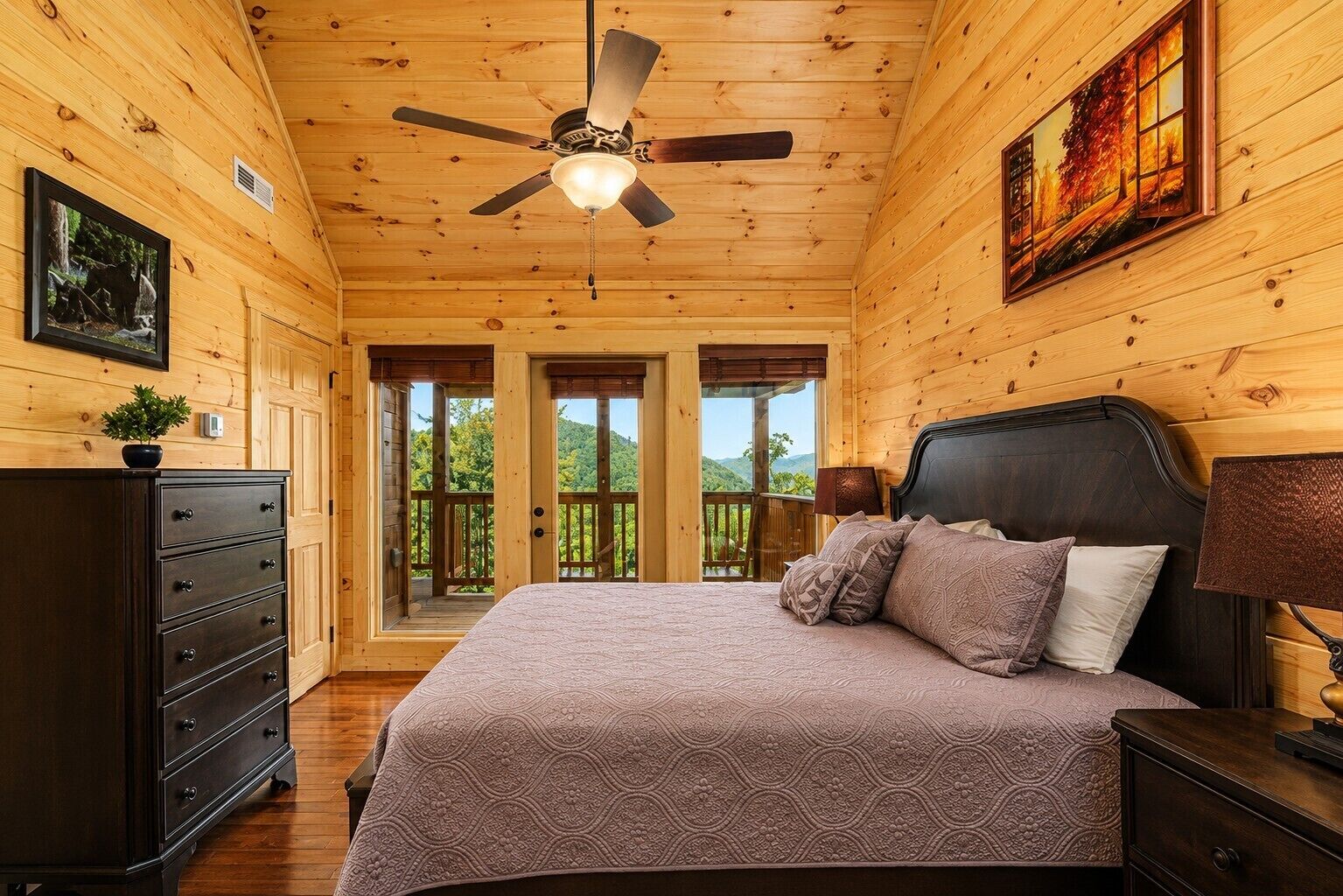 A wooden cabin bedroom with a bed, dresser, ceiling fan, and a painting on the wall.
