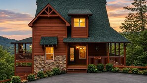 A two-story log cabin with a stone foundation, a wrap-around porch, and a view of the surrounding forest. - Cabin + FREE Resort Amenities | Pigeon Forge (Pigeon Forge)