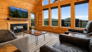 A wooden interior with a fireplace, a television, and a large window overlooking a deck. - Luxury Cabin w/ All-Inclusive Resort Amenities (Sevierville)
