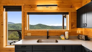 A wooden cabin kitchen with a large window, a sink, cabinets, and a toaster. - Luxury Cabin w/ All-Inclusive Resort Amenities (Sevierville)