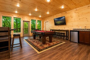 A wooden interior with a pool table, bar area, and a refrigerator.