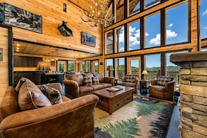 A spacious living room with leather sofas, a coffee table, and a stone fireplace. Large windows offer a view of the outdoors.