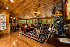 A cabin interior with wooden walls, a fireplace, a sofa, a patterned armchair, and a dining area.