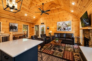 A log cabin interior with a dining area, a living room with a leather sofa, a fireplace, and a ceiling fan.