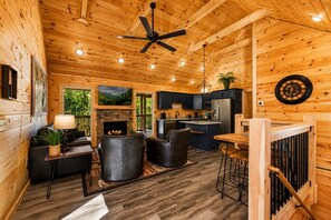 A cabin interior with a fireplace, seating area, kitchen, and a ceiling fan.