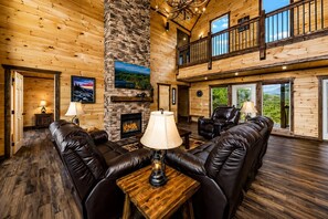 A spacious living room with a stone fireplace, leather sofas, and a large window offering a view of the outdoors.