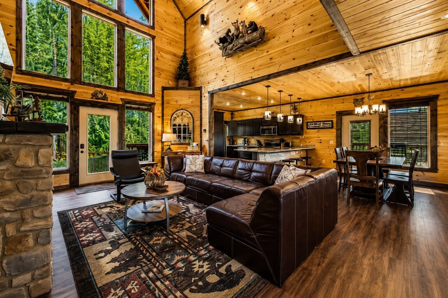 A spacious living room with a leather sofa, a stone fireplace, and a large window overlooking a forest.