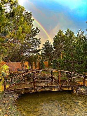 A rustic wooden bridge over a clear stream, surrounded by pine trees and a stone structure in the background. - Lepista Garden (Mengen)