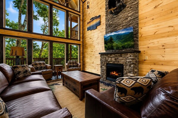 A cozy living room with leather sofas, a fireplace, and large windows overlooking a forest.
