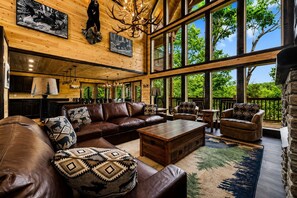 A spacious living room with a large wooden ceiling, leather furniture, and a view of the outdoors through large windows. - Luxury Cabin w/ All-Inclusive Resort Amenities (Sevierville)