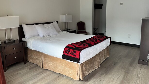 A neatly made bed with a white comforter and a red and black blanket, two bedside lamps, a wooden nightstand, a chair, and a wardrobe in a hotel room. - The Driftwood Lodge (Juneau)