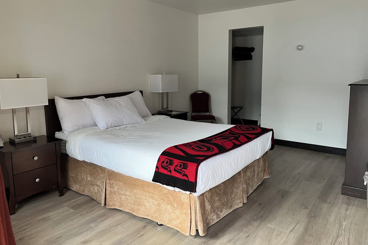 Cama bem arrumada com edredom branco e manta preta e vermelha, duas luminárias de cabeceira, mesa de cabeceira de madeira, uma cadeira e um guarda-roupa em um quarto de hotel.