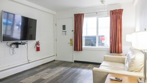 A hotel room with a flat-screen TV mounted on the wall, a sofa, a small table with a remote control, and a window with curtains. - The Driftwood Lodge (Juneau)