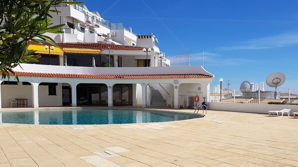 A poolside area with a clear blue sky, a building with white walls and red-tiled roofs, and a satellite dish on the roof.