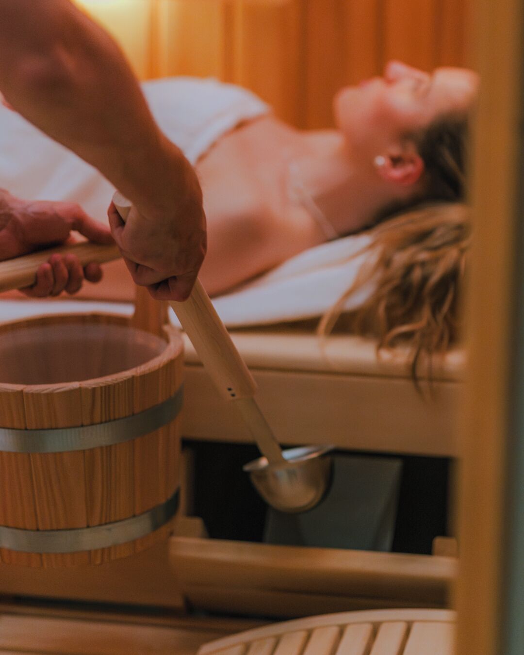 Eine Person rührt mit einem Holzlöffel in einem großen Holzbehälter, vermutlich einer Hot Tub, in einem Raum, in dem sich eine andere Person befindet.