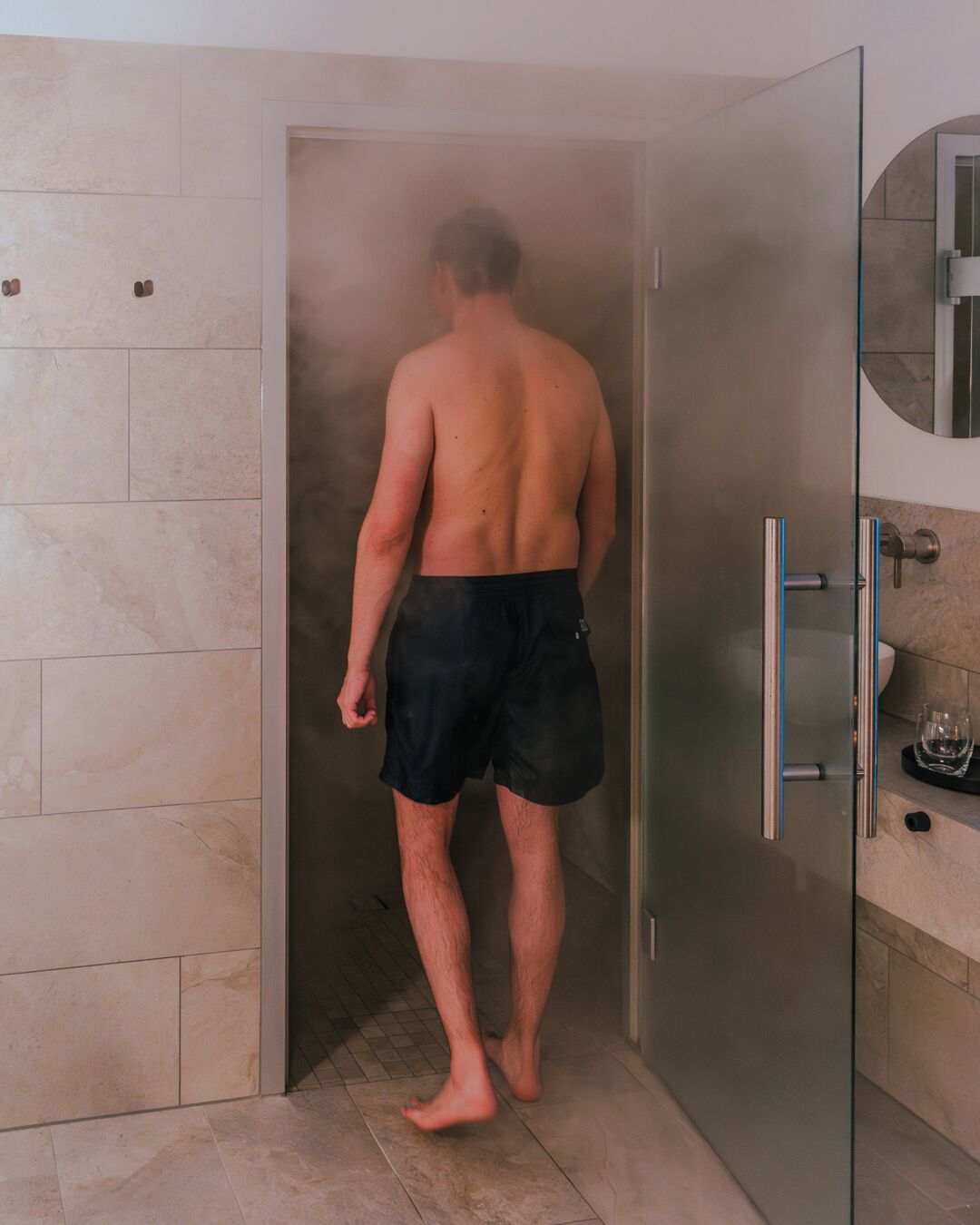 A man in black shorts walking into a steam shower with a glass door.