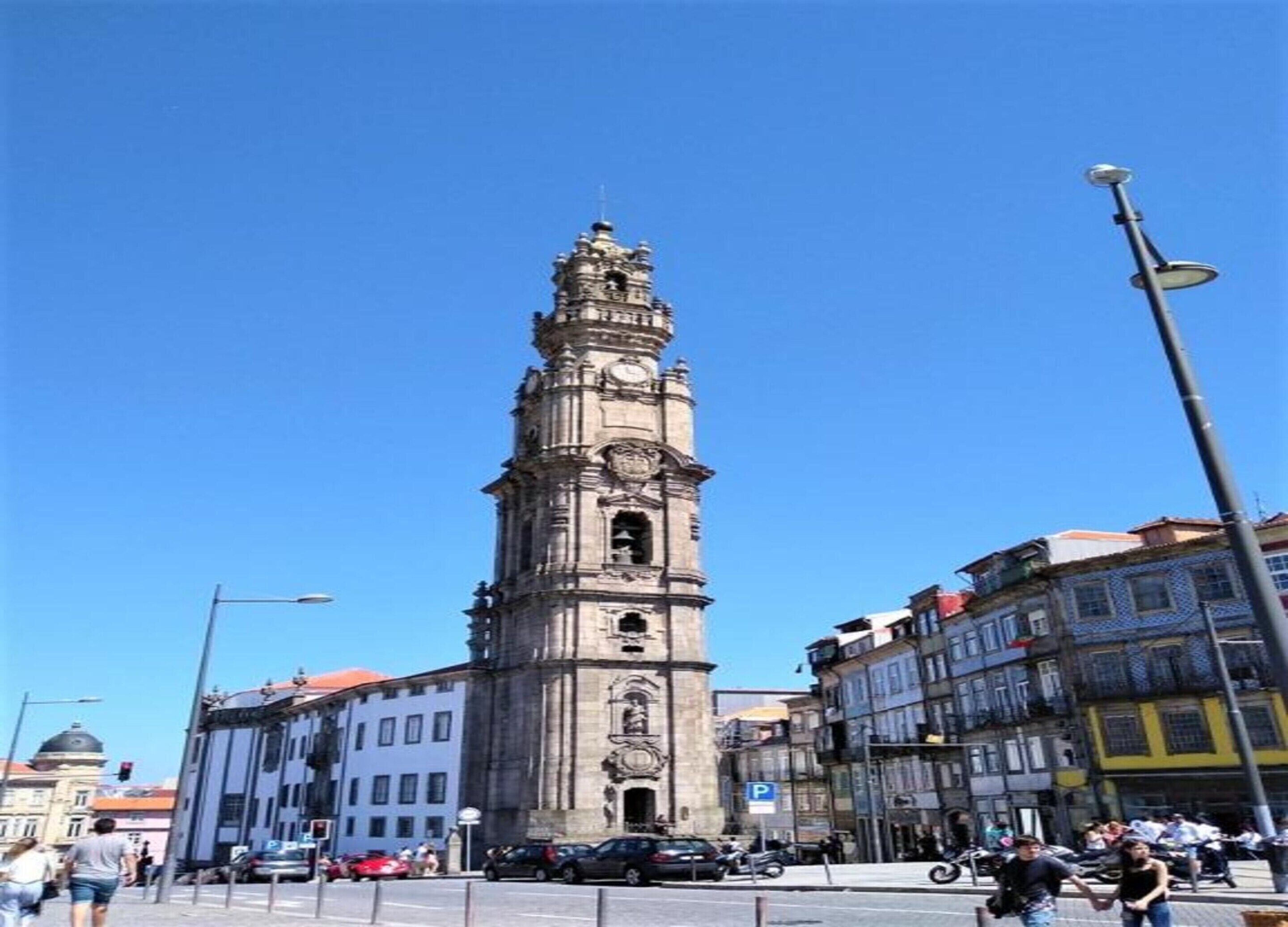 Una torre del reloj histórica con arquitectura elaborada, rodeada de edificios urbanos y un cielo azul despejado.