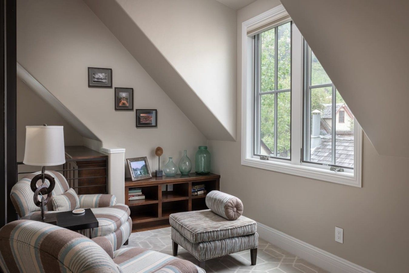 A cozy loft bedroom with a sloped ceiling, a large window, a built-in bookshelf, and a striped armchair.
