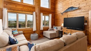 Un intérieur de chalet chaleureux avec une grande télévision, des murs en bois, un canapé et une vue par les fenêtres.