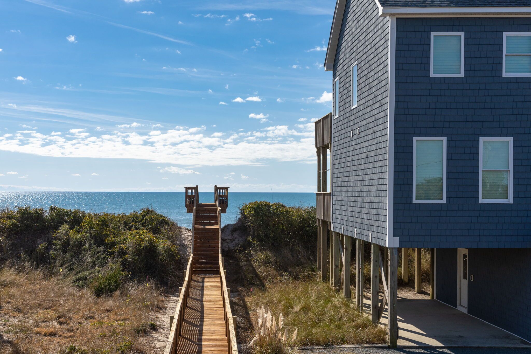 Een houten looppad leidt naar een donkerblauwe woning met een balkon, met uitzicht op een strand.
