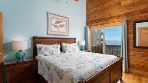 Une chambre avec un plafond en bois, un grand lit recouvert d’une literie à motifs floraux, deux tables de chevet avec des lampes et une fenêtre donnant sur un beau paysage.
