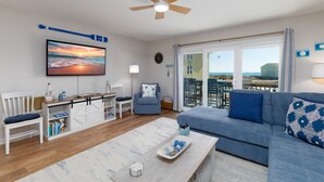 A modern living room with a sofa, a TV, a ceiling fan, and a view of the sea through large windows. - Blue Oar- Beach, Pool, and Sun in Surf City! (Surf City)