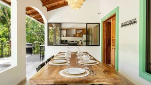 A dining area with a long wooden table set for a meal, a kitchen visible through a glass partition, and a sign reading 'BEACH TOWELS' on the wall. - Selva Blanca, Jungle Retreat (Playa Hermosa)
