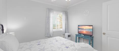 Un dormitorio con cama, ventilador de techo, televisor y ventana con cortinas.