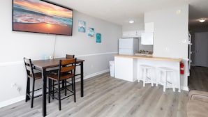 A modern kitchen with a wooden dining table and chairs, a white refrigerator, and a bar area with stools. - Sun of a Beach- Your Ultimate Beachy Getaway (North Topsail Beach)