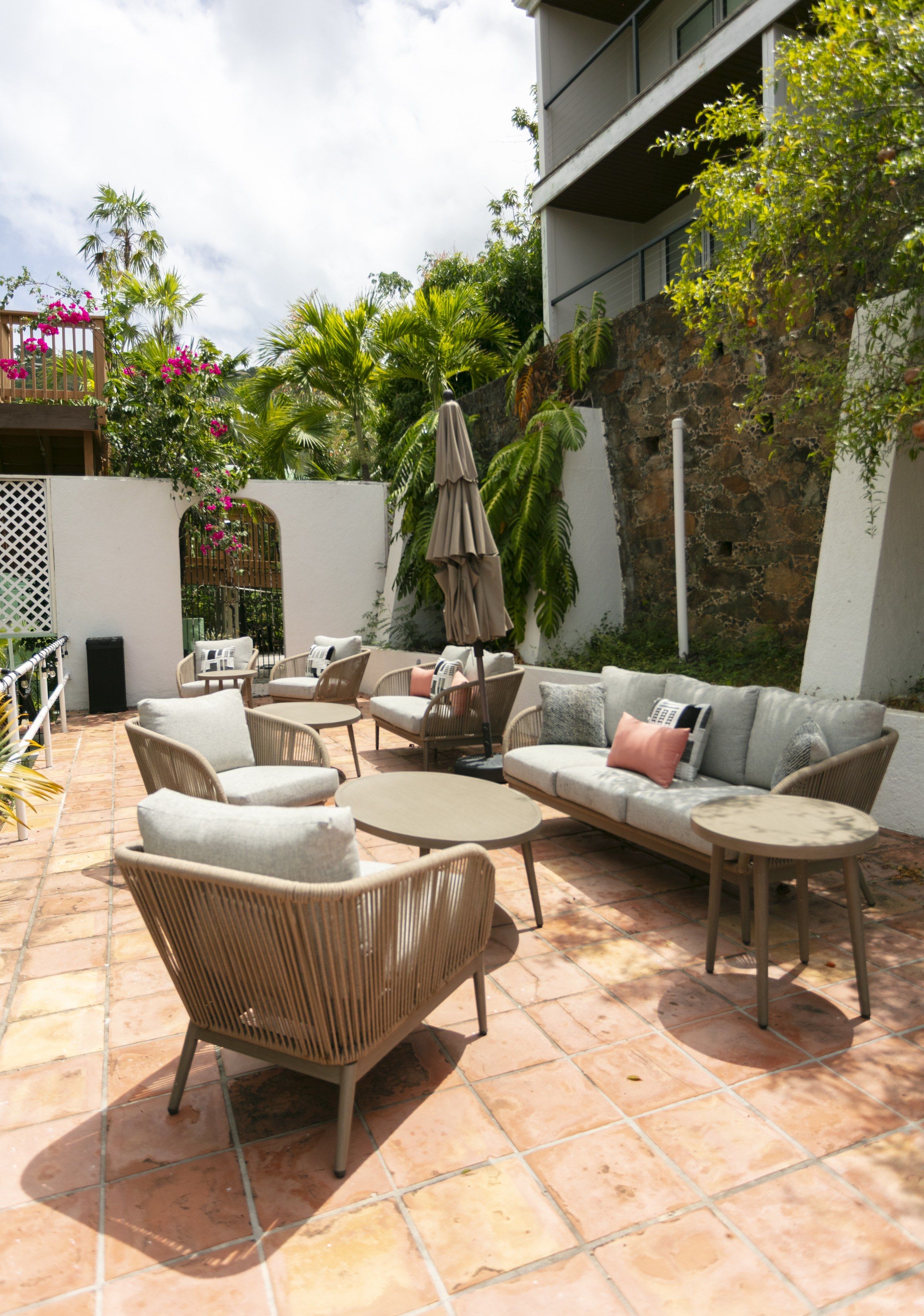 An outdoor patio with wicker furniture, a stone wall, and lush greenery.