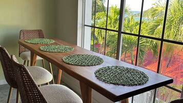 A dining area with a long wooden table, wicker chairs, and a large window with a view of greenery and a distant landscape.