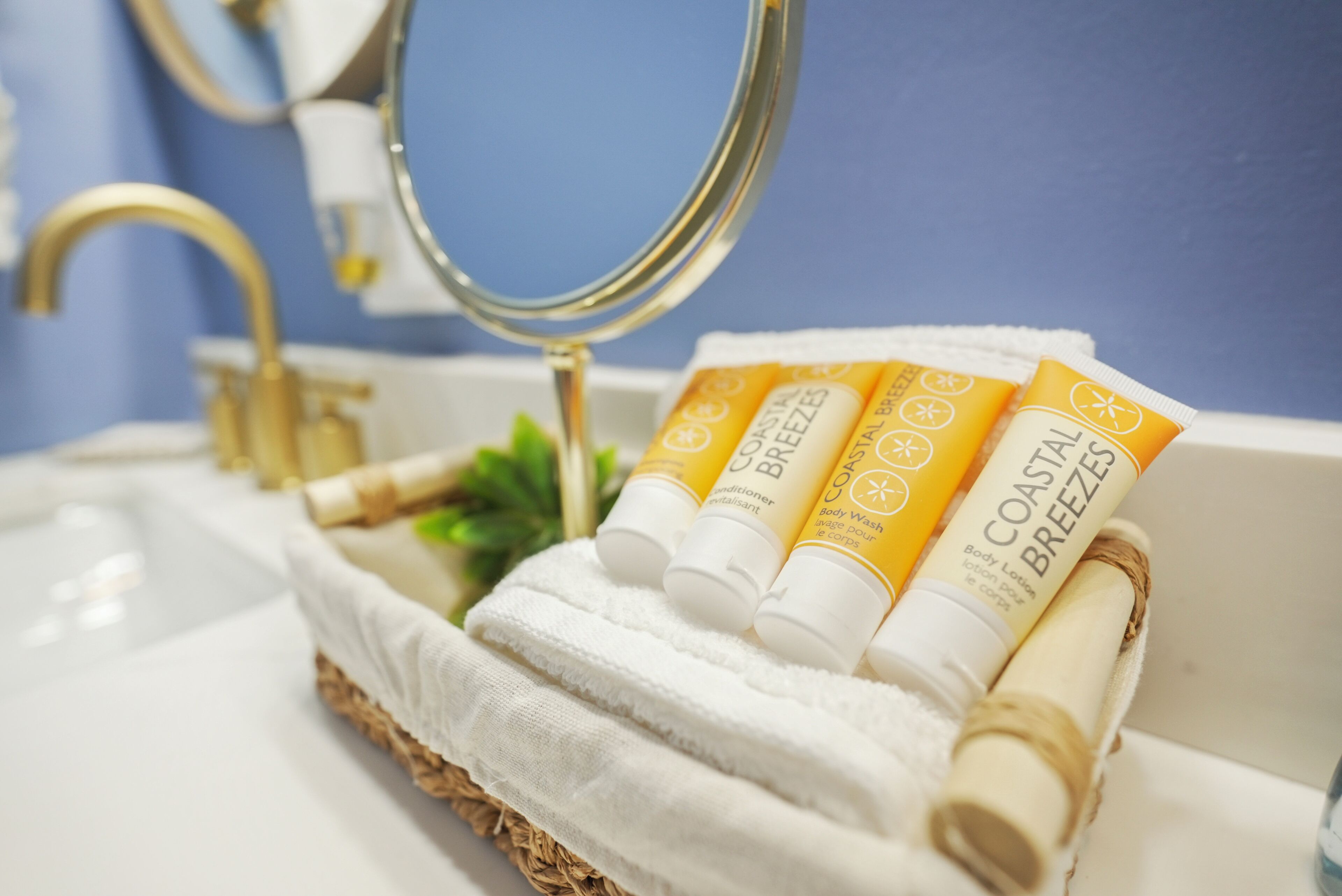 A bathroom with a white sink, a gold faucet, and a mirror. There are three tubes of body lotion and a rolled-up towel on a white cloth.