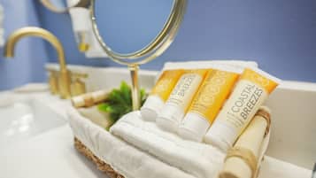 A bathroom with a white sink, a gold faucet, and a mirror. There are three tubes of body lotion and a rolled-up towel on a white cloth.