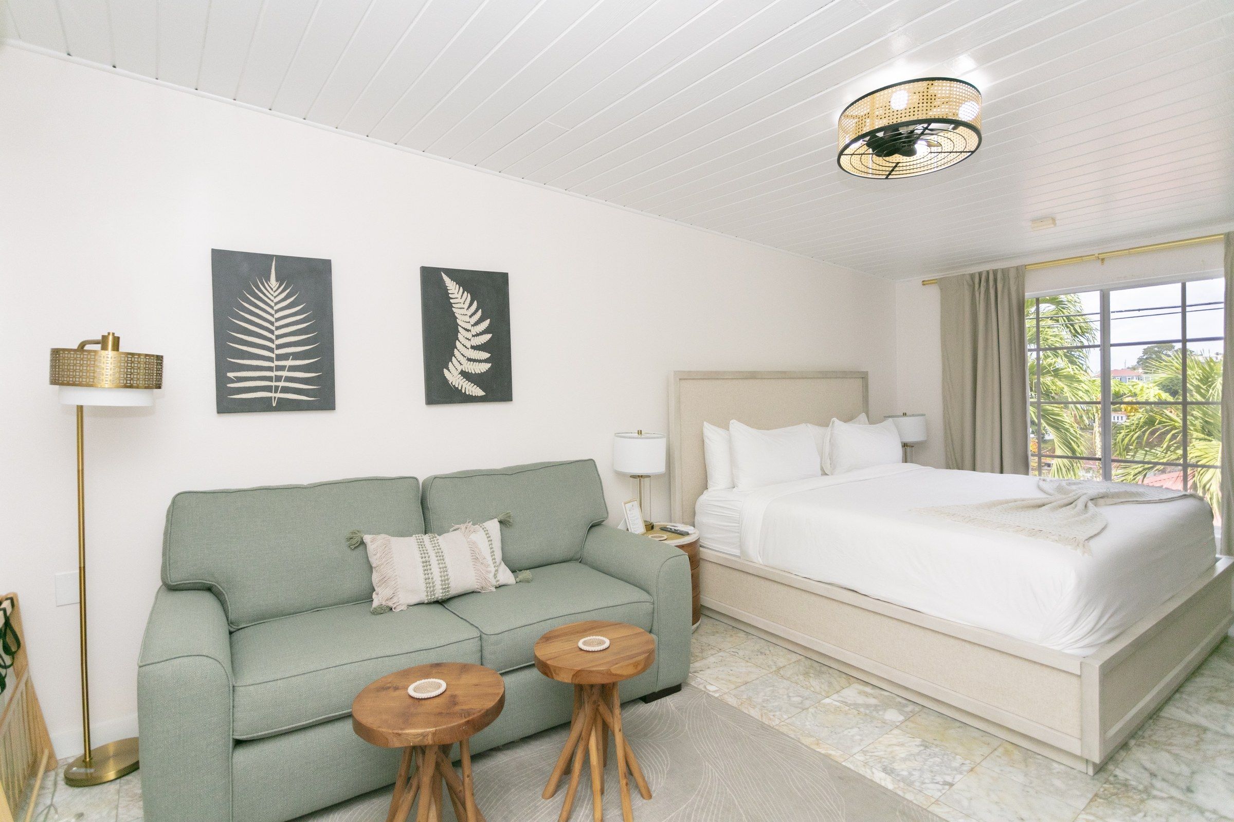 A hotel room with a bed, a green sofa, two wooden side tables, and two wall-mounted fern artwork.