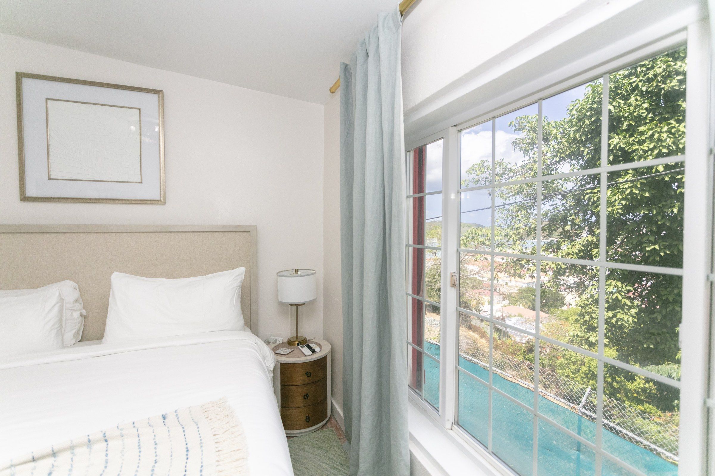 A hotel room with a bed, a bedside table with a lamp, a framed picture on the wall, and a window with a view of greenery.