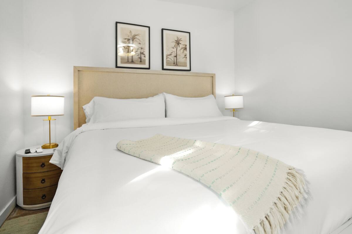A neatly made bed with white linens, a beige throw blanket, and two framed pictures on the wall above.
