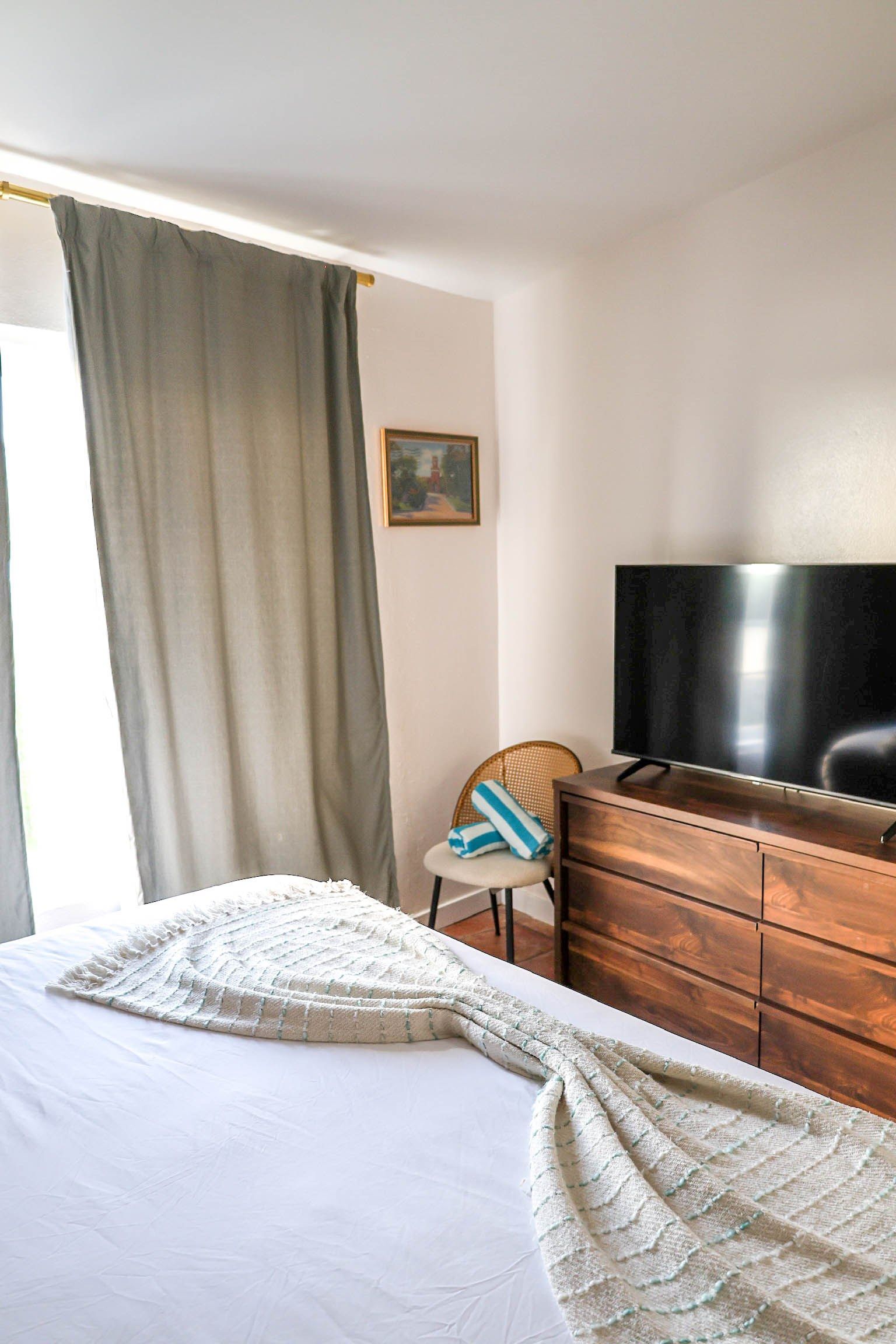 Une chambre d’hôtel avec un lit, une commode en bois, une télévision, une chaise et un tableau accroché au mur.