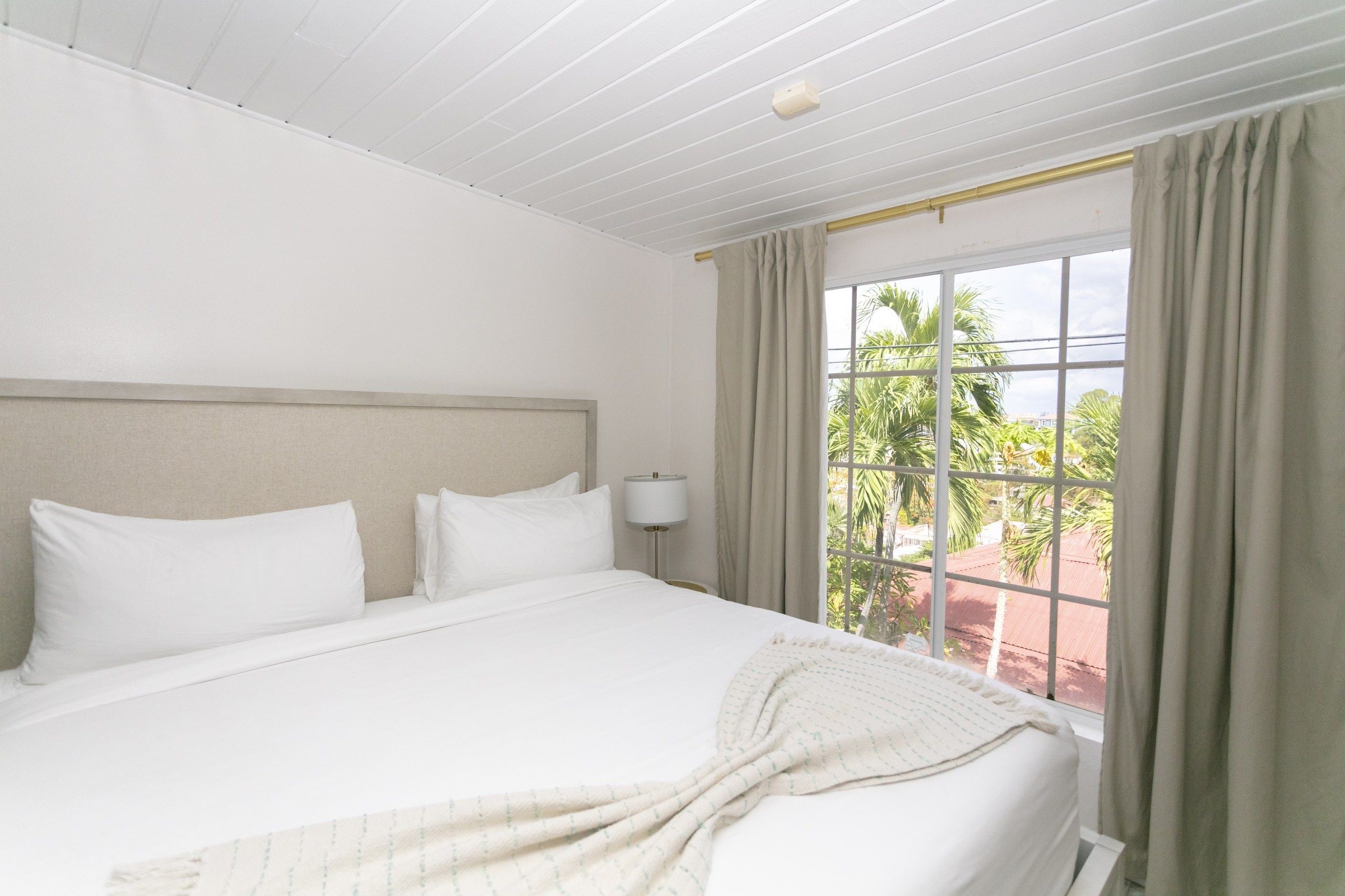 Habitación de hotel con una cama grande, una mesita de noche, una lámpara y un ventanal con cortinas.