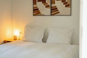 Un lit bien fait, avec une literie blanche, une lampe de chevet et une œuvre d’art abstraite accrochée au mur.