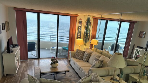 A living room with a sofa, a coffee table, and a TV. There is a balcony with a view of the ocean. - Oceanfront Sand Dollar Condo 601 (Daytona Beach Shores)