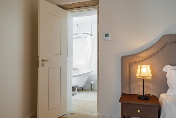 Une chambre d’hôtel avec une porte blanche, une table de chevet avec une lampe, un lit et une vue sur une salle de bain équipée d’une baignoire et d’une douche.