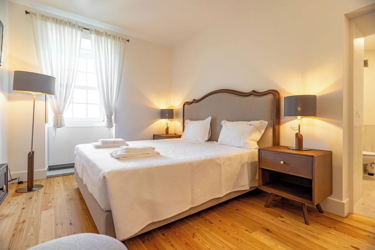 Une chambre d’hôtel avec un grand lit, deux tables de chevet avec des lampes, un plancher en bois et une fenêtre avec des rideaux.