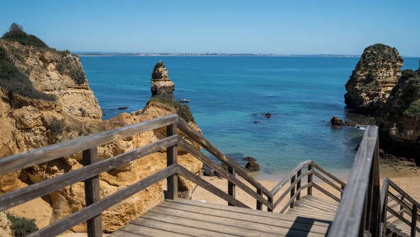 Un escalier en bois mène à une plage où l’eau est d’un bleu cristallin et où se dressent des falaises rocheuses.